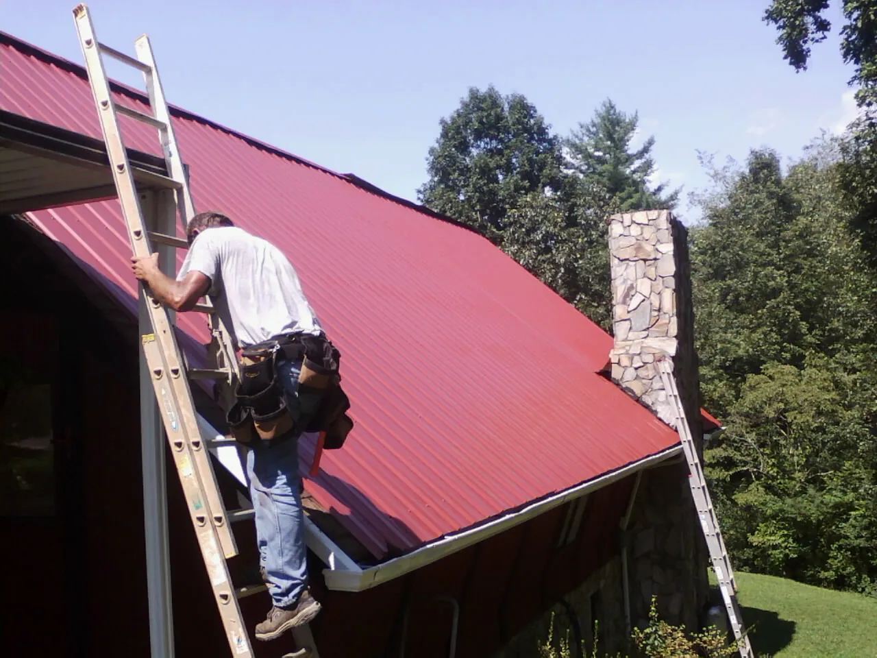 Roofer on a ladder beside a red metal roof and stone chimney — Soffit & Fascia Repair by Roofing Brothers in Ewell
